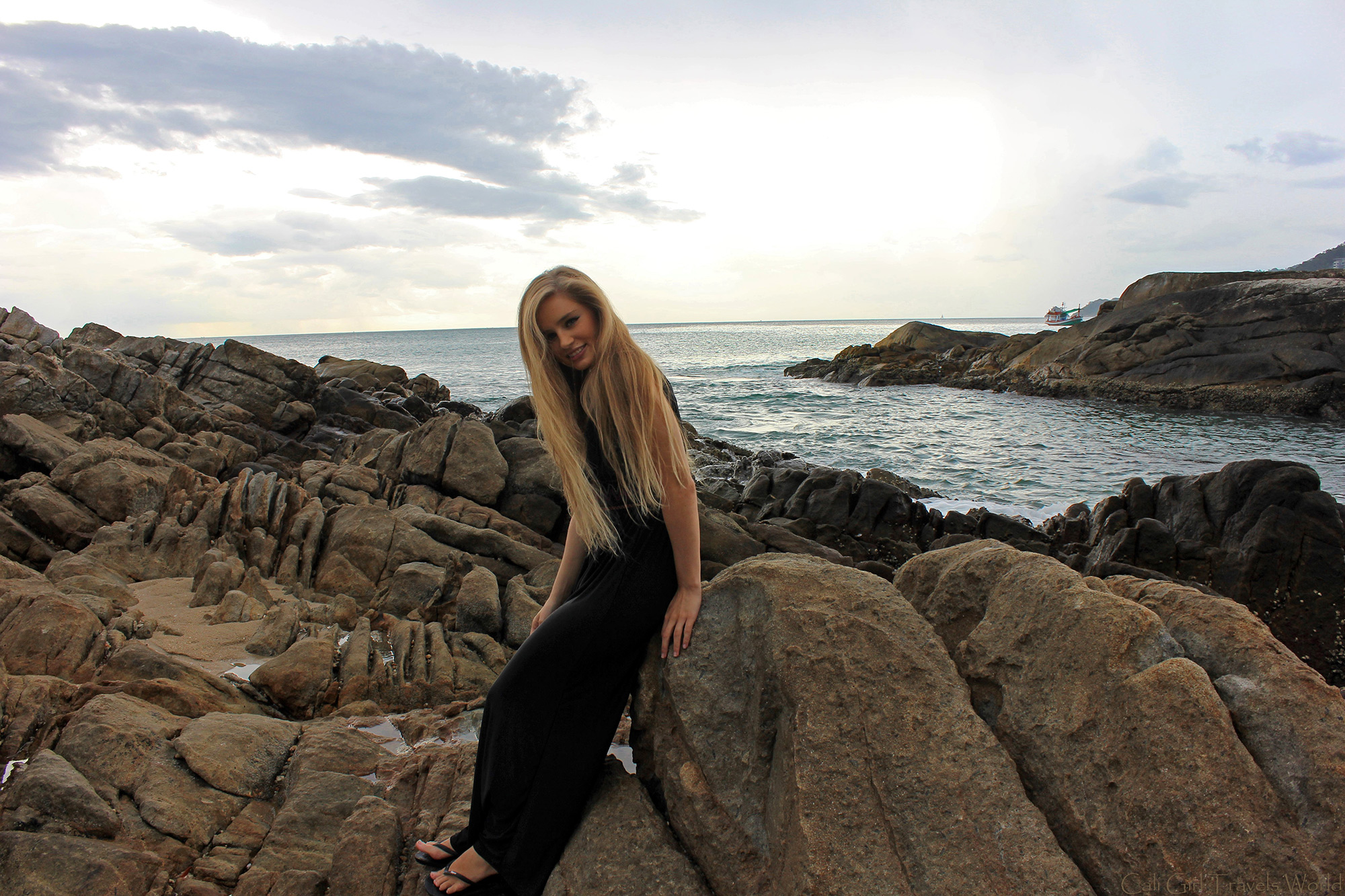 Blonde female travel blogger in long black dress at rocky beach smiling.